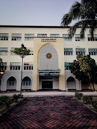 A tall, multi-story building with a prominent entrance decorated in an architectural style featuring geometric patterns. The sign 'Dentistry' is displayed above the entrance. The building is surrounded by a few trees and neatly trimmed bushes. A tiled pathway leads up to the entrance, and a traffic cone is placed near the doorway.