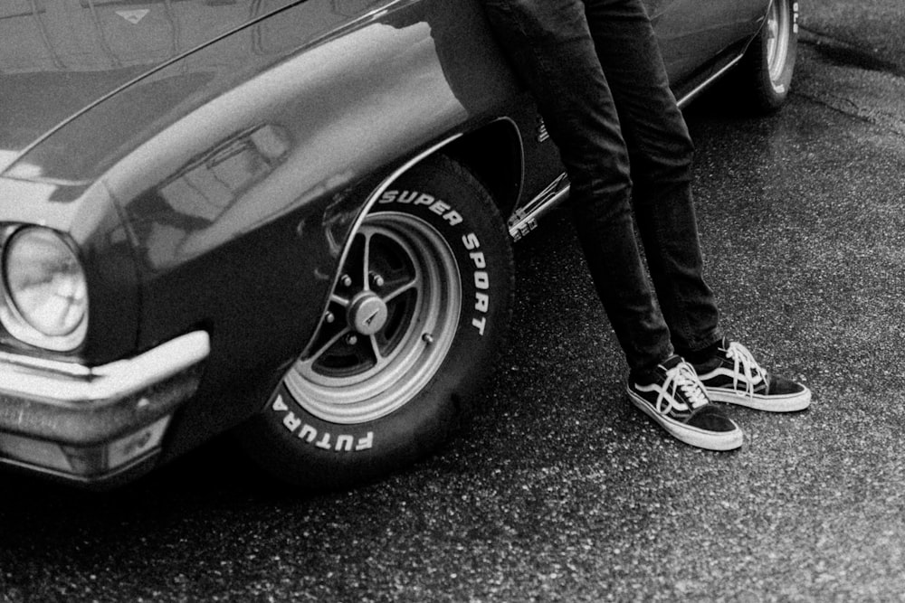grayscale photo of person in black pants and sneakers standing beside car