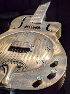 A resonator guitar with a metallic body featuring intricate perforations and a distinctive finish. The image highlights the guitar's round resonator cover and the knobs for volume and tone control, with strings and tuning pegs extending out of view. The lighting accentuates the texture and details of the guitar's surface.