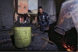 A man wearing a dark jacket works near a large metal container that appears to be used for heating or burning, evident from the glowing embers and scattered sparks. The scene is set in an outdoor workshop area with weathered walls and scattered building materials. A rusty yellow barrel is prominently placed to the side, and in the background, the facade of an old building and some parked motorcycles are visible.