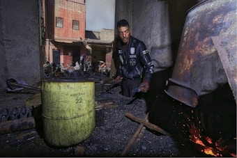 A man wearing a dark jacket works near a large metal container that appears to be used for heating or burning, evident from the glowing embers and scattered sparks. The scene is set in an outdoor workshop area with weathered walls and scattered building materials. A rusty yellow barrel is prominently placed to the side, and in the background, the facade of an old building and some parked motorcycles are visible.