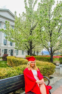 A person wearing a red graduation cap and gown sits on a bench in a park-like setting. Surrounding them are blooming trees, well-manicured hedges, and a path leading to a stately building with pillars and large windows.