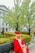 A person wearing a red graduation cap and gown sits on a bench in a park-like setting. Surrounding them are blooming trees, well-manicured hedges, and a path leading to a stately building with pillars and large windows.
