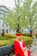 A person wearing a red graduation cap and gown sits on a bench in a park-like setting. Surrounding them are blooming trees, well-manicured hedges, and a path leading to a stately building with pillars and large windows.