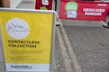 A yellow sign displays information about a contactless collection service, mentioning social distancing and sanitizing guidelines. It is set on the pavement near a parking area. Behind the sign, there is a red banner indicating a dedicated parking spot for a park and pick-up service.