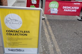A yellow sign displays information about a contactless collection service, mentioning social distancing and sanitizing guidelines. It is set on the pavement near a parking area. Behind the sign, there is a red banner indicating a dedicated parking spot for a park and pick-up service.