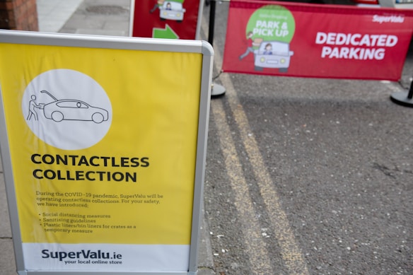 A yellow sign displays information about a contactless collection service, mentioning social distancing and sanitizing guidelines. It is set on the pavement near a parking area. Behind the sign, there is a red banner indicating a dedicated parking spot for a park and pick-up service.