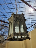 An ornate black lantern, possibly made of metal, featuring intricate gothic-style designs, hangs suspended from a chain. The background displays a metal grid roof structure with clear blue skies and sunlight streaming in.