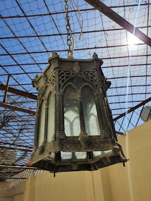 An ornate black lantern, possibly made of metal, featuring intricate gothic-style designs, hangs suspended from a chain. The background displays a metal grid roof structure with clear blue skies and sunlight streaming in.