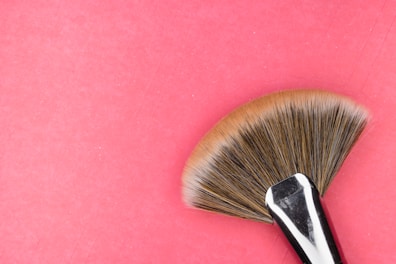 A makeup fan brush with a polished metal handle and soft, fanned out bristles is placed against a pink background.