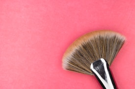 A makeup fan brush with a polished metal handle and soft, fanned out bristles is placed against a pink background.