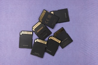 A group of seven black SD cards is scattered on a textured purple surface. The cards are arranged with their gold contacts facing upwards, showing a mix of logos and manufacturing details.