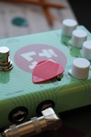 A close-up of a guitar effects pedal featuring a light blue and green patterned design with white knobs. There is a red guitar pick resting on top of the pedal, and a metallic audio jack is plugged into one of the ports.