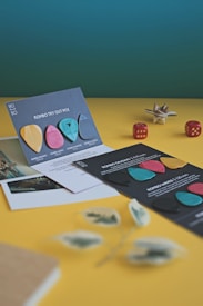 A neatly arranged display of colorful guitar picks on top of dark informational cards is set against a vibrant yellow surface. Some are upright and others are slightly tilted, showing different designs and textures. Two red dice and a decorative star sculpture are placed around the scene. Blurred in the foreground are hints of a plant and some documents.