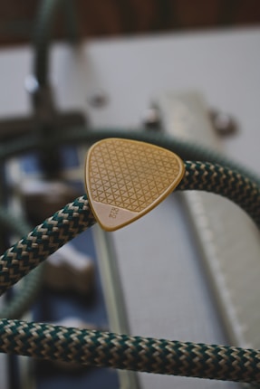 A close-up image of an acoustic guitar's strings, showcasing a triangular thumb pick nestled between them. The pick has a textured surface featuring a grid pattern and a small printed text. The strings are intertwined with green and beige patterns, contributing to a sense of detail and complexity against a blurred background of the guitar's body.