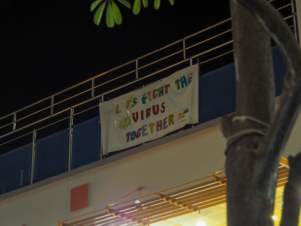 A banner hangs on the side of a building under dim lighting. The banner has the message 'Let's Fight The Virus Together' in colorful letters with a small illustration of a virus.