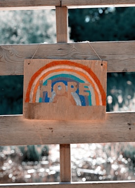 A wooden sign is hanging from a wooden fence, painted with a rainbow design featuring red, blue, orange, and light blue arcs. The word 'HOPE' is written in the center of the rainbow. The background is blurry with round light reflections, suggesting an outdoor setting with sunlight filtering through foliage.