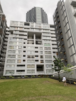 A modern high-rise building with a unique architectural design featuring multiple balconies and large windows. In the foreground, a grassy area with a few trees is present, and a person is walking across the lawn while holding a child's hand. The sky is overcast, giving the scene a calm and muted atmosphere.