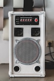 A white audio amplifier with a prominent speaker on the front, featuring a metal grille with a circular design. Above the speaker are red control knobs for various audio settings such as mic volume, echo, bass, treble, and balance. The device is rectangular and placed on a flat surface with several acoustic panels behind it.