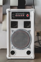 A white audio amplifier with a prominent speaker on the front, featuring a metal grille with a circular design. Above the speaker are red control knobs for various audio settings such as mic volume, echo, bass, treble, and balance. The device is rectangular and placed on a flat surface with several acoustic panels behind it.