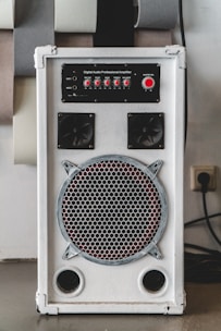 A white audio amplifier with a prominent speaker on the front, featuring a metal grille with a circular design. Above the speaker are red control knobs for various audio settings such as mic volume, echo, bass, treble, and balance. The device is rectangular and placed on a flat surface with several acoustic panels behind it.