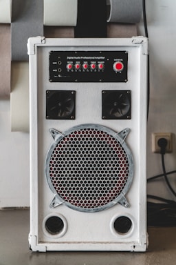 A white audio amplifier with a prominent speaker on the front, featuring a metal grille with a circular design. Above the speaker are red control knobs for various audio settings such as mic volume, echo, bass, treble, and balance. The device is rectangular and placed on a flat surface with several acoustic panels behind it.