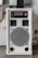 A white audio amplifier with a prominent speaker on the front, featuring a metal grille with a circular design. Above the speaker are red control knobs for various audio settings such as mic volume, echo, bass, treble, and balance. The device is rectangular and placed on a flat surface with several acoustic panels behind it.