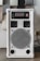 A white audio amplifier with a prominent speaker on the front, featuring a metal grille with a circular design. Above the speaker are red control knobs for various audio settings such as mic volume, echo, bass, treble, and balance. The device is rectangular and placed on a flat surface with several acoustic panels behind it.