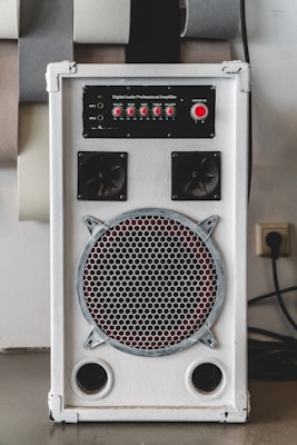 A white audio amplifier with a prominent speaker on the front, featuring a metal grille with a circular design. Above the speaker are red control knobs for various audio settings such as mic volume, echo, bass, treble, and balance. The device is rectangular and placed on a flat surface with several acoustic panels behind it.