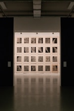 A gallery setting with a grid of framed artworks on the wall, each depicting close-up, detailed textures resembling lunar surfaces or celestial bodies. The lighting is subdued, creating a focused and contemplative atmosphere.