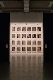 A gallery setting with a grid of framed artworks on the wall, each depicting close-up, detailed textures resembling lunar surfaces or celestial bodies. The lighting is subdued, creating a focused and contemplative atmosphere.
