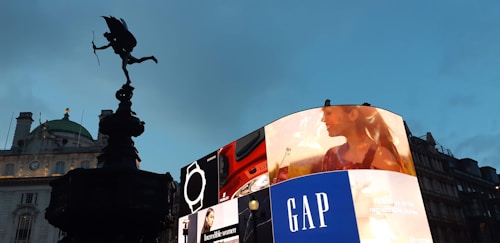 A city scene featuring a prominent digital billboard displaying multiple advertisements, including one for GAP and an image of a woman. To the left, a silhouetted statue of a winged figure stands against a background of historic architecture and a cloudy evening sky.