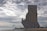 A large stone monument depicting figures standing on a ship, with intricate carvings and statues. The structure stands near a waterfront, with a cloudy sky overhead. Cars and people can be seen near the base of the monument.