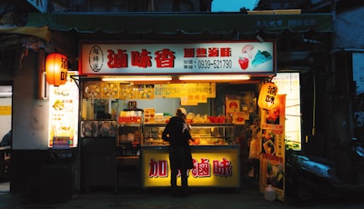A brightly lit street food stall with a variety of snacks displayed. One person stands in front, likely ordering or examining the items. The stall features colorful signage with large text and images of food, illuminated by warm lights. Nearby, a scooter is parked, and surrounding buildings are dimly lit, creating a cozy and inviting atmosphere.