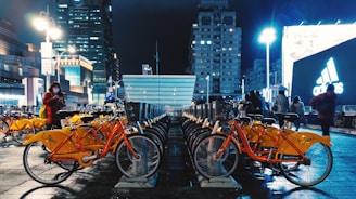 A bustling urban scene at night featuring a row of bright orange rental bicycles neatly parked on a wet pavement. In the background, tall illuminated buildings and billboards, including an Adidas advertisement, create a vibrant city atmosphere. Several people can be seen walking and one person is using a smartphone.