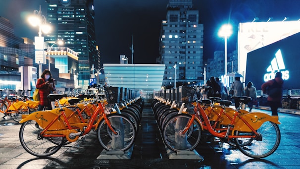A bustling urban scene at night featuring a row of bright orange rental bicycles neatly parked on a wet pavement. In the background, tall illuminated buildings and billboards, including an Adidas advertisement, create a vibrant city atmosphere. Several people can be seen walking and one person is using a smartphone.