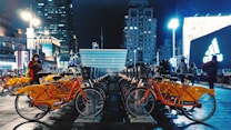 A bustling urban scene at night featuring a row of bright orange rental bicycles neatly parked on a wet pavement. In the background, tall illuminated buildings and billboards, including an Adidas advertisement, create a vibrant city atmosphere. Several people can be seen walking and one person is using a smartphone.