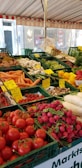 A vibrant outdoor market stall filled with a variety of fresh vegetables. The display includes tomatoes, radishes, carrots, cucumbers, mushrooms, and bell peppers, all neatly organized in green crates. Handwritten price labels are visible throughout, giving a sense of a bustling farmers' market.