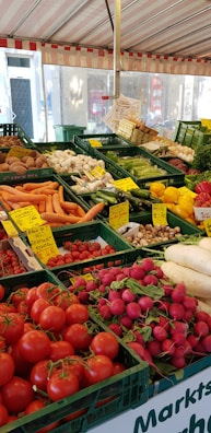 A vibrant outdoor market stall filled with a variety of fresh vegetables. The display includes tomatoes, radishes, carrots, cucumbers, mushrooms, and bell peppers, all neatly organized in green crates. Handwritten price labels are visible throughout, giving a sense of a bustling farmers' market.