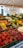 A vibrant outdoor market stall filled with a variety of fresh vegetables. The display includes tomatoes, radishes, carrots, cucumbers, mushrooms, and bell peppers, all neatly organized in green crates. Handwritten price labels are visible throughout, giving a sense of a bustling farmers' market.
