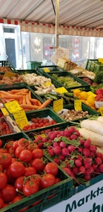 A vibrant outdoor market stall filled with a variety of fresh vegetables. The display includes tomatoes, radishes, carrots, cucumbers, mushrooms, and bell peppers, all neatly organized in green crates. Handwritten price labels are visible throughout, giving a sense of a bustling farmers' market.