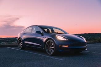 A sleek, blue electric car is parked in an empty parking lot during sunset. The car has a modern design with a smooth and aerodynamic shape. The sky features a gradient of orange and pink hues as the sun sets on the horizon, casting a gentle light on the vehicle.