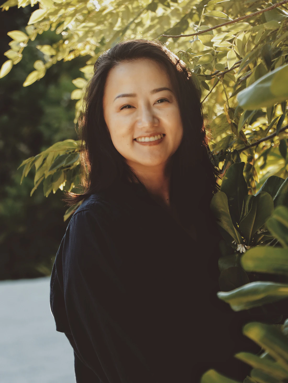 A smiling woman in her fifties standing among green leaves in soft light