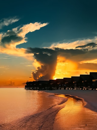 Overwater bungalows line a serene beach at sunset, with large, dramatic clouds illuminated by the orange and yellow hues of the setting sun reflecting on the calm sea.