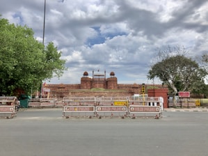 A historic red sandstone fort stands prominently with three domes and various decorative elements. The sky above is filled with dramatic clouds, casting a moody atmosphere over the scene. Several barricades marked with 'Delhi Police' block the foreground, and there are lush green trees on either side of the frame.