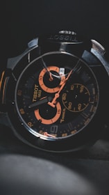 A close-up of a wristwatch with a black and orange color scheme, featuring intricate details on the face, multiple dials, and visible branding.