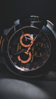 A close-up of a wristwatch with a black and orange color scheme, featuring intricate details on the face, multiple dials, and visible branding.