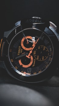 A close-up of a wristwatch with a black and orange color scheme, featuring intricate details on the face, multiple dials, and visible branding.