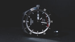 A sleek wristwatch with a black dial displaying the brand name and a date window. The watch features luminous hands, minute markers around the outer rim, and a red accent at the tip of the second hand. The bezel is marked with numbers in increments of ten.