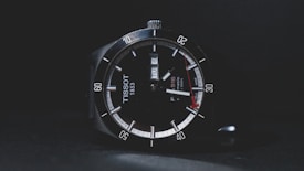 A sleek wristwatch with a black dial displaying the brand name and a date window. The watch features luminous hands, minute markers around the outer rim, and a red accent at the tip of the second hand. The bezel is marked with numbers in increments of ten.